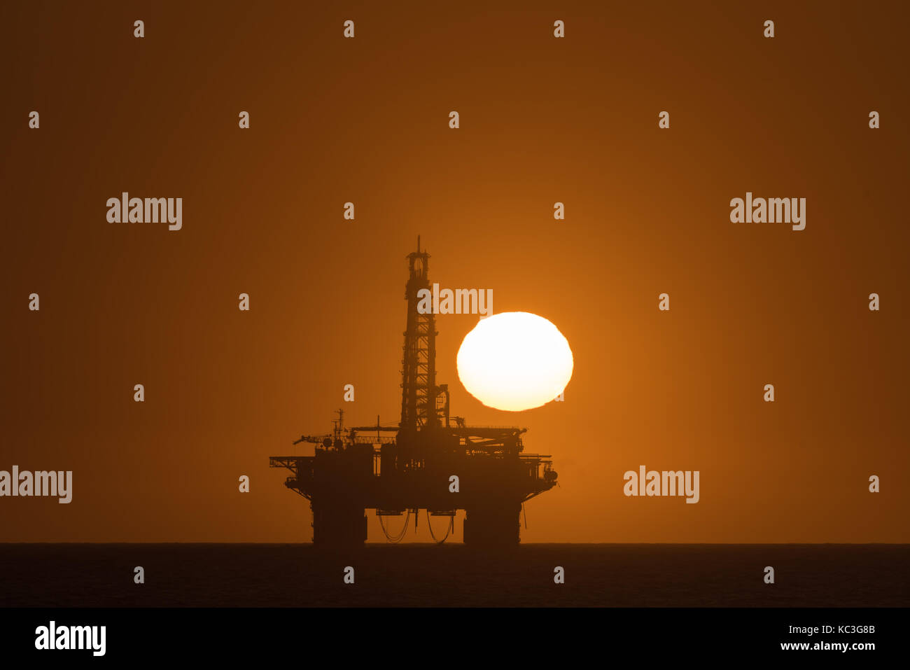 The sun is setting over an oil drilling platform anchored in the Atlantic Ocean at Longbeach in the Namib Desert of Namibia Stock Photo