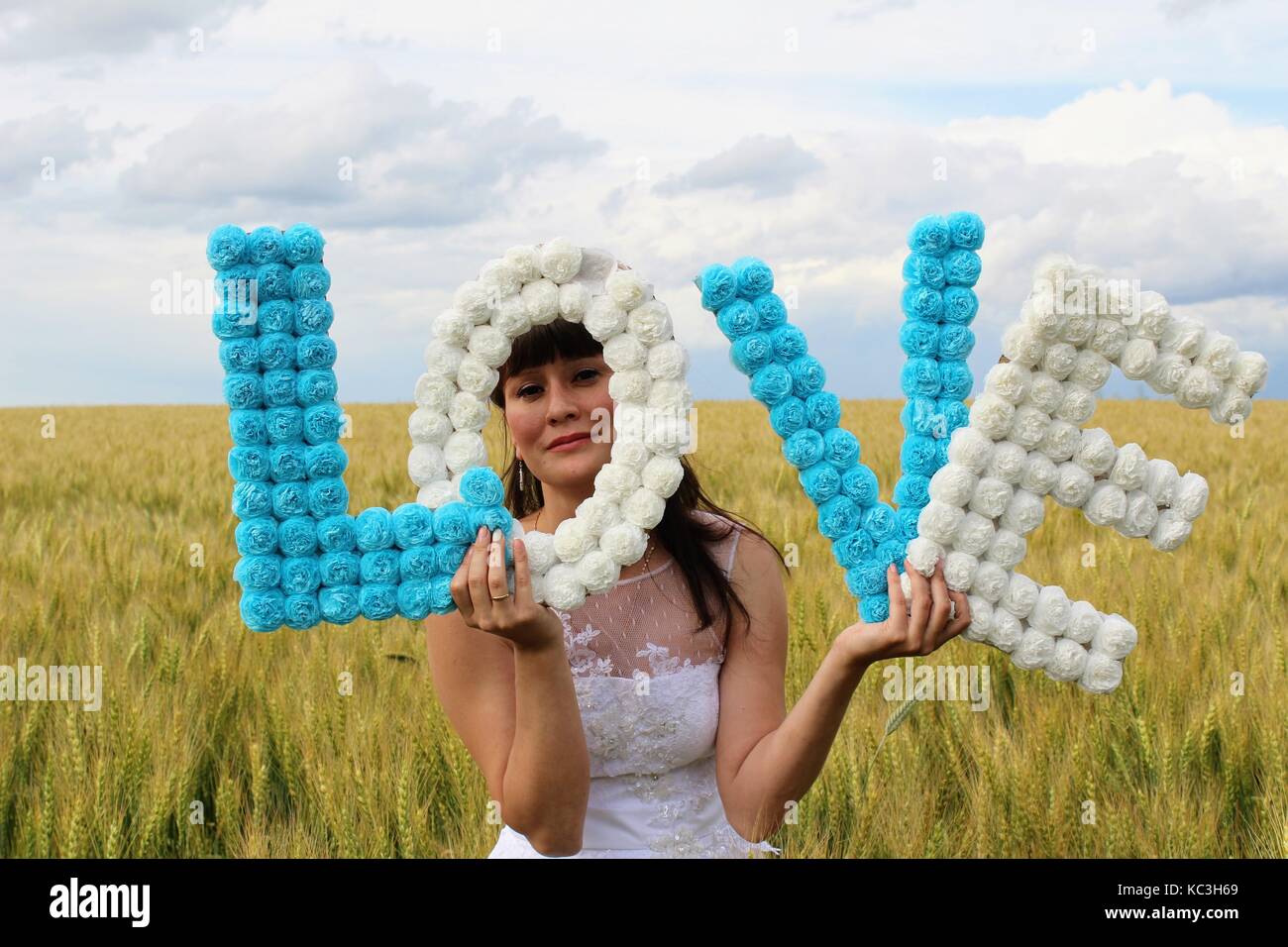 A girl in a wedding dress is holding the letters 'LOVE' standing against a wheat field and blue sky. Stock Photo