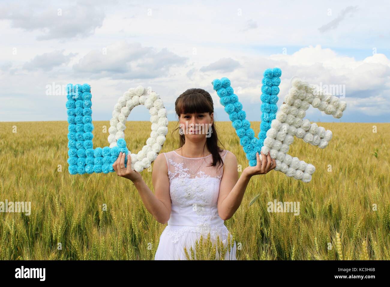 A girl in a wedding dress is holding the letters 'LOVE' standing against a wheat field and blue sky. Stock Photo