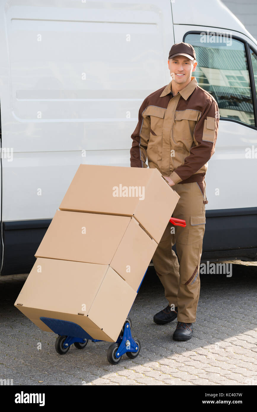 Portrait of confident delivery man pushing parcels on handtruck against van Stock Photo