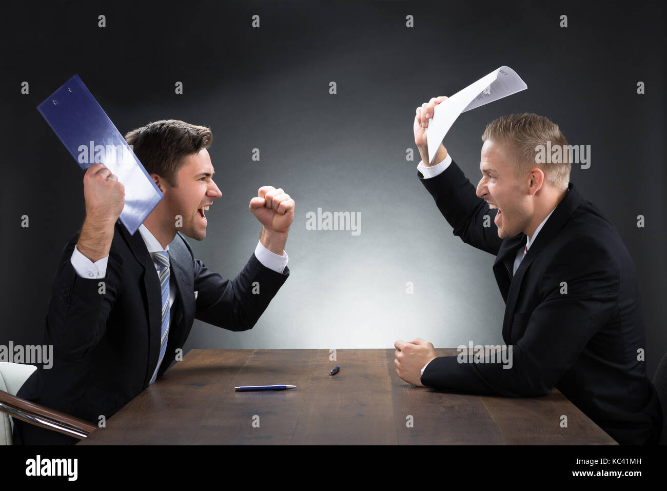 Aggressive young businessmen arguing at wooden desk against gray background Stock Photo