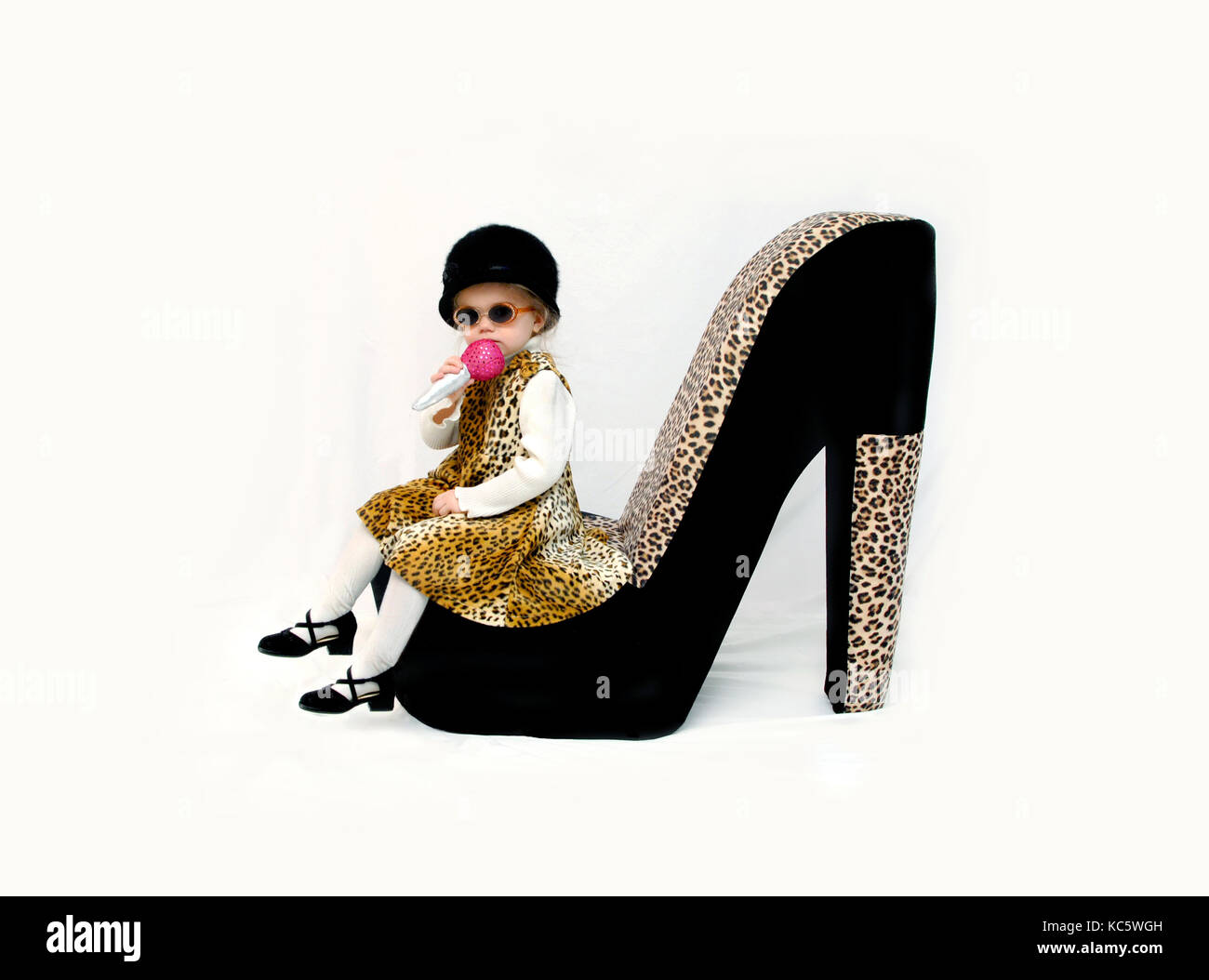 Little girl, wearing a leopard print dress and black furry hat, sings into a pretend microphone.  She is sitting on an animal print giant shoe. Stock Photo