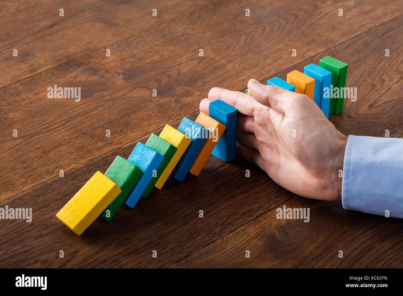 Person Hand Stopping Falling Dominoes From Continuous Toppled On Wooden Desk. Business Risk Concept Stock Photo