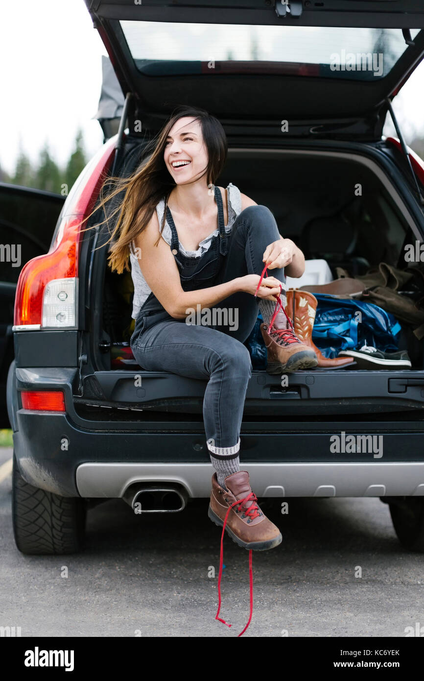 Smiling woman sitting in car trunk and tying hiking shoes Stock Photo