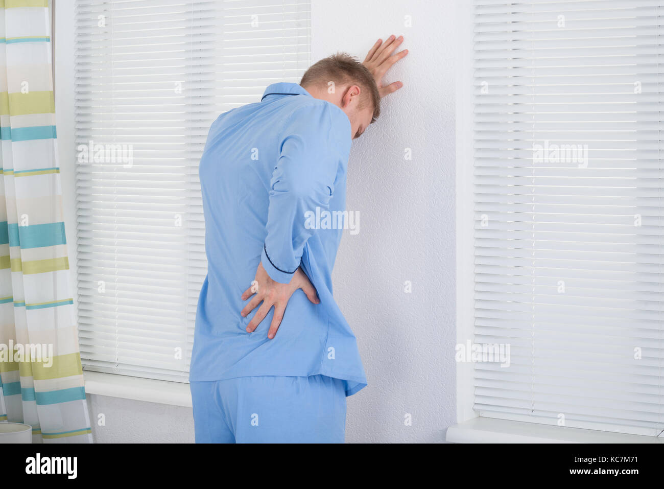 Man Suffering From Back Ache Standing Against Wall Stock Photo