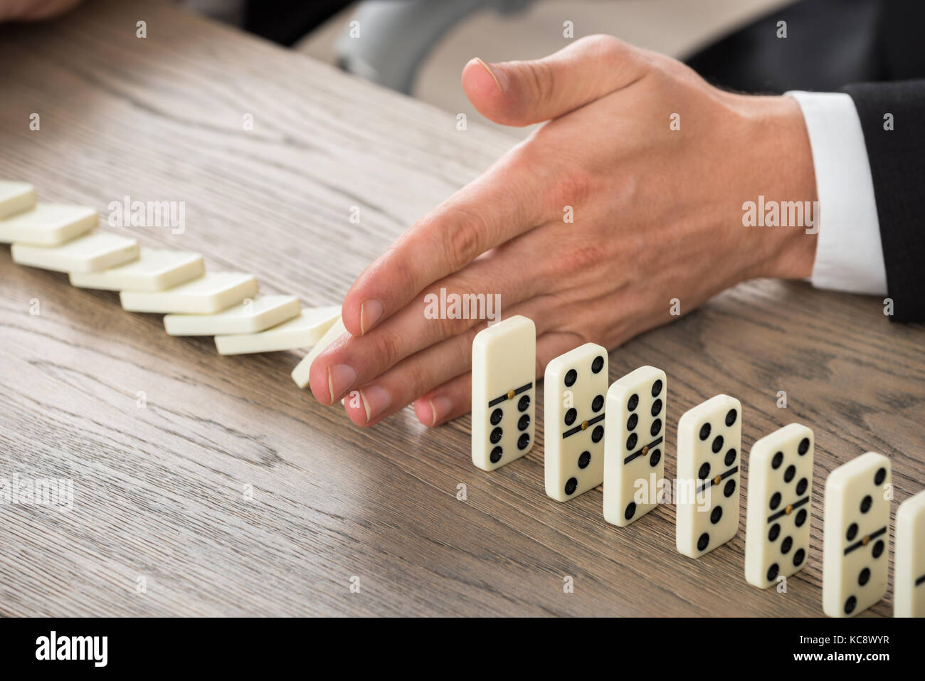 Close-up Of A Businessman Hand Stopping Dominoes From Falling On A Desk In Office Stock Photo