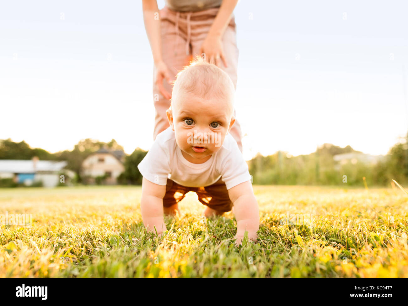 Cute baby boy with his unrecognizable mother crawling outside on the green grass. Stock Photo