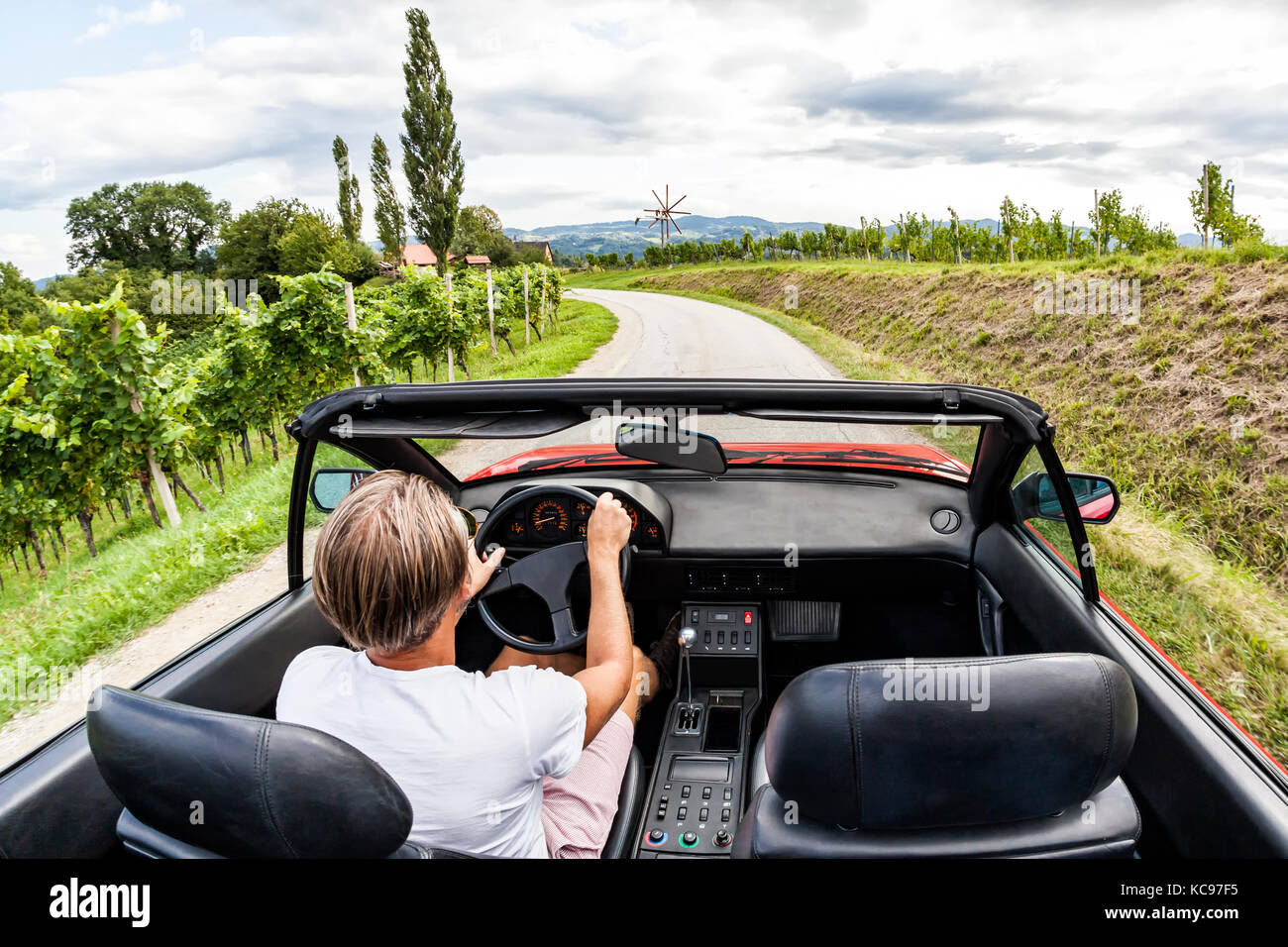 Young man driving with red vintage sports car through the vineyard on south styrian vine route in Austria. Windmill called Klapotetz in front. Stock Photo
