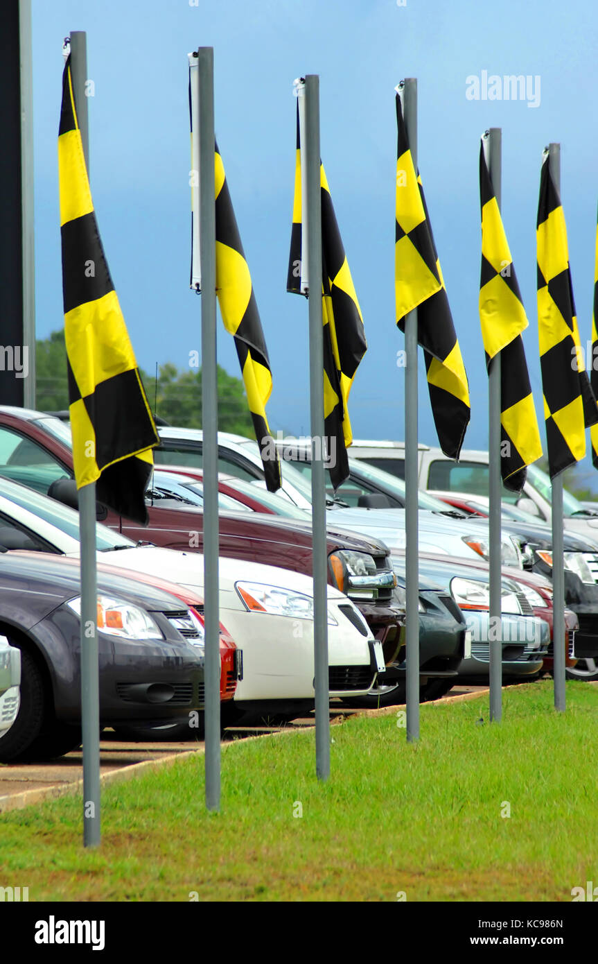 Automobiles line up against the curb at a used car lot.  Checkered flags flutter in the breeze to attract customers to stop and browse. Stock Photo