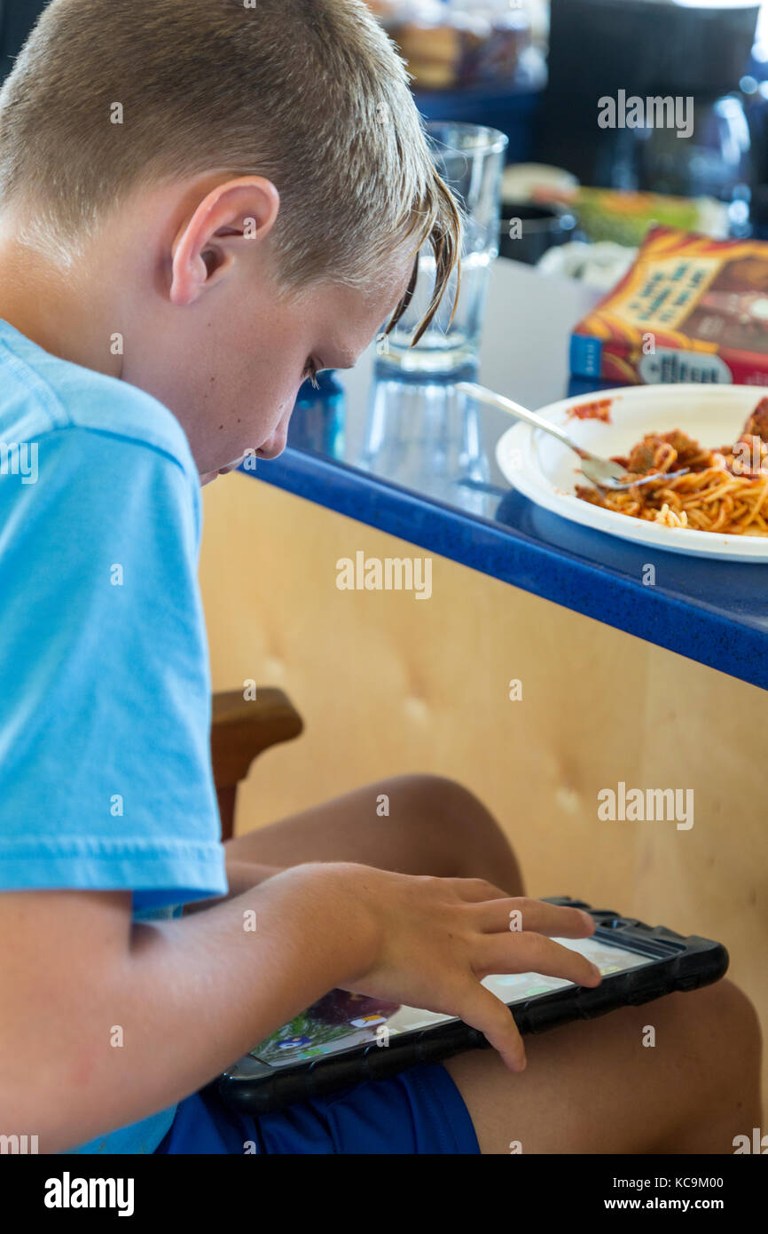 Avon, Outer Banks, North Carolina, USA.  Young Boy Eating Lunch while Playing with Mobile Gaming Device. Stock Photo