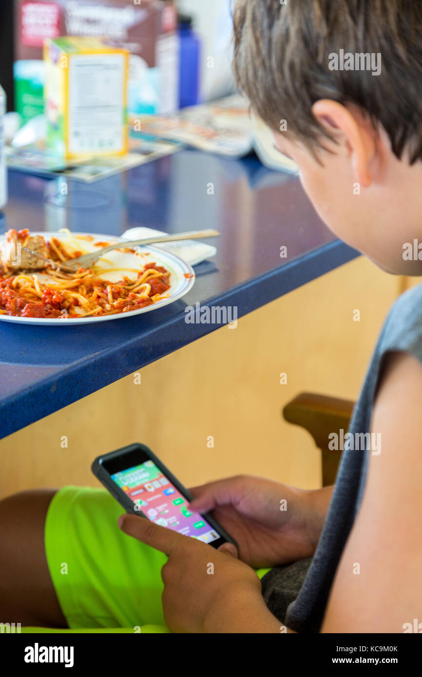 Avon, Outer Banks, North Carolina, USA.  Young Boy Eating Lunch while Playing with Mobile Gaming Device. Stock Photo