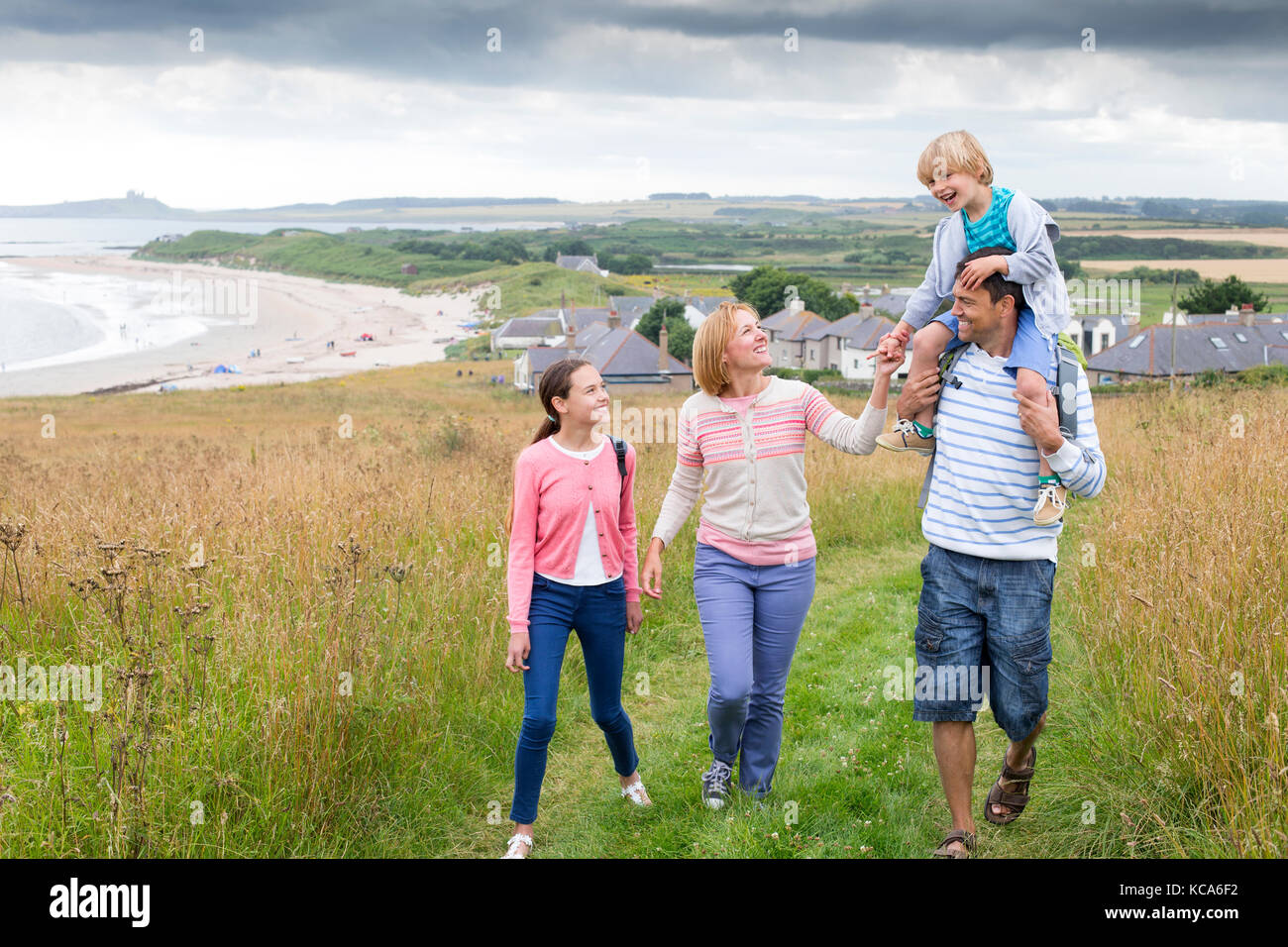 A family of four are walking on the sand dunes together. The son is sitting on his fathers shoulders. They all look happy and are smiling. Stock Photo