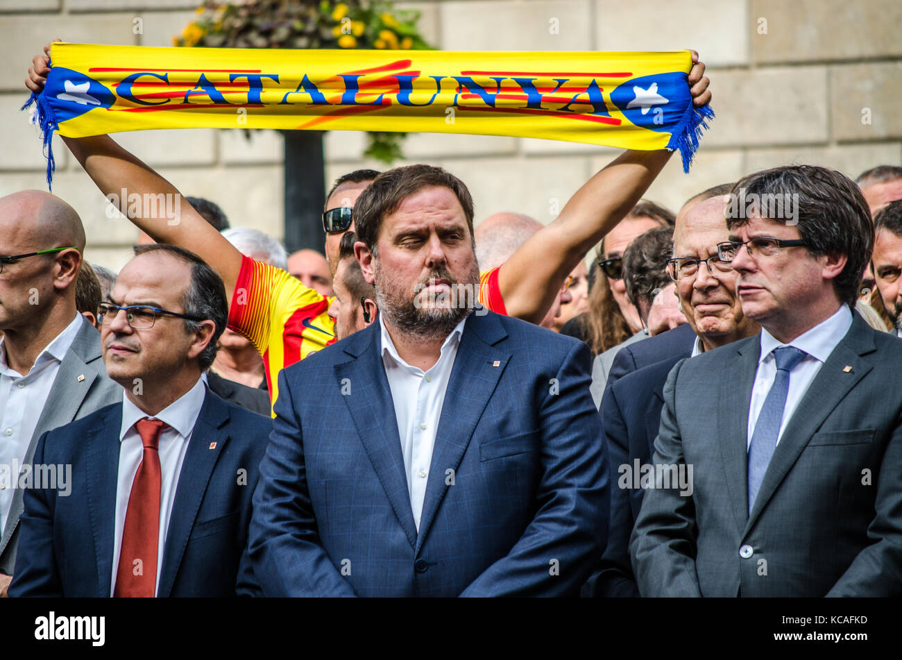 Barcelona, Spain. 3rd Oct, 2017. Jordi Turull, Oriol Junqueras, Carles Puigdemont, (left to right) are seen during the minute of silence, while a man raising in the air a Catalonia scarf. The Catalan Government and the city of Barcelona gathered for a minute of silence as a sign of rejection against Police brutality, in the San Jauma Plaza, headquarters of both administrations. Stock Photo