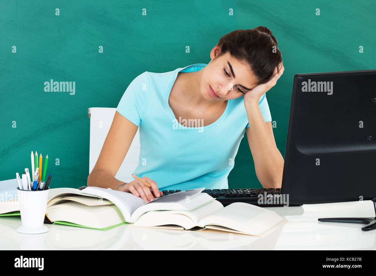 Stressed Young Woman Studying On Desk With Pile Of Books And Computer Desktop In Classroom Stock Photo