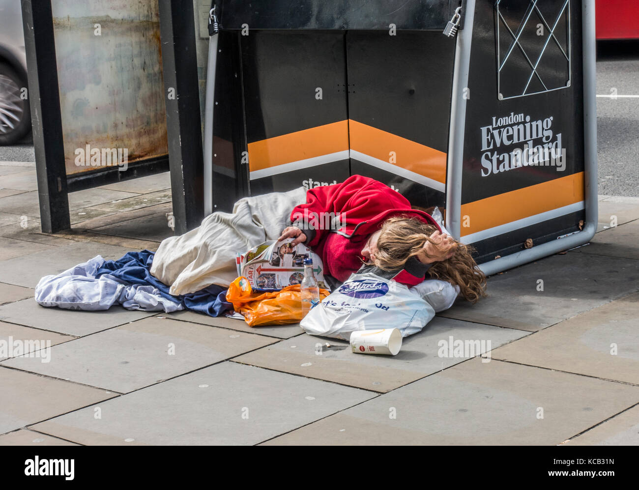 Rough sleeper / homeless person, reading a magazine next to an Evening Standard newspaper stand, lying down on the pavement in London, England, UK. Stock Photo