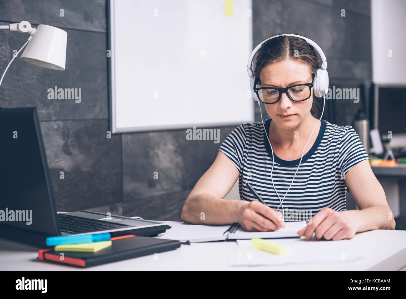 Woman wearing eyeglasses writing notes and listening music at the office Stock Photo