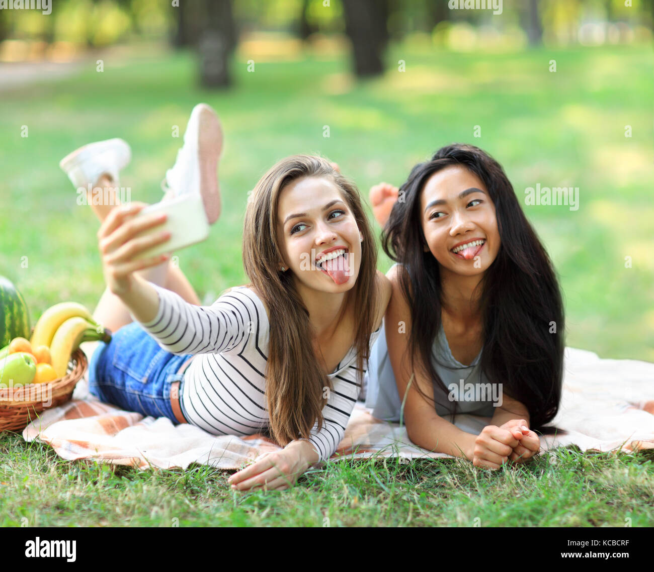 Caucasian and Asian young woman doing selfie and showing tongues. Funny girl friends having fun in free time in sunny park. International women friend Stock Photo