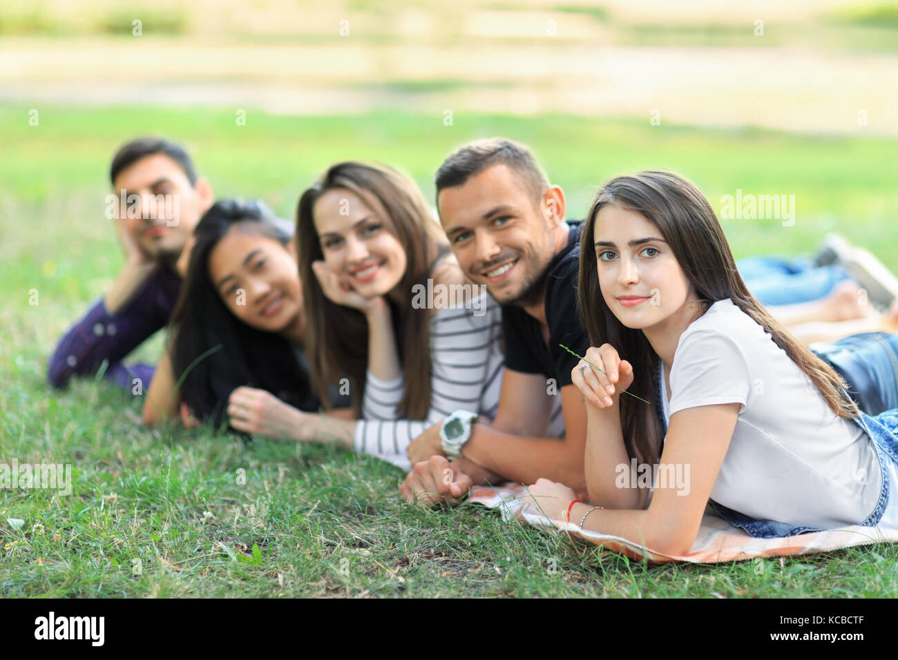 Group of five friends lying in row in park. Smiling young people with different ethnicity having fun outdoors. Focus on woman. Carefree students leisu Stock Photo