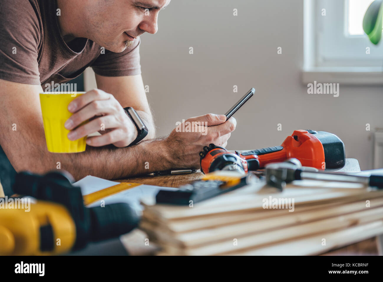 Man using smart phone and holding yellow cup of coffee in front of table with tools at home Stock Photo
