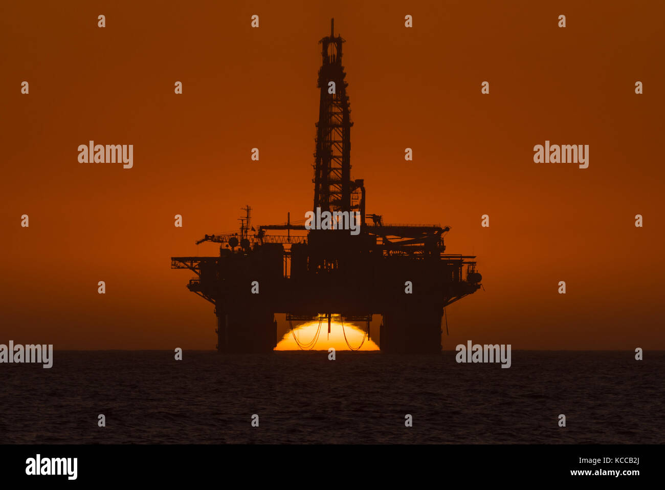 The sun is setting behind an oil drilling platform anchored in the Atlantic Ocean at Longbeach in the Namib Desert of Namibia Stock Photo