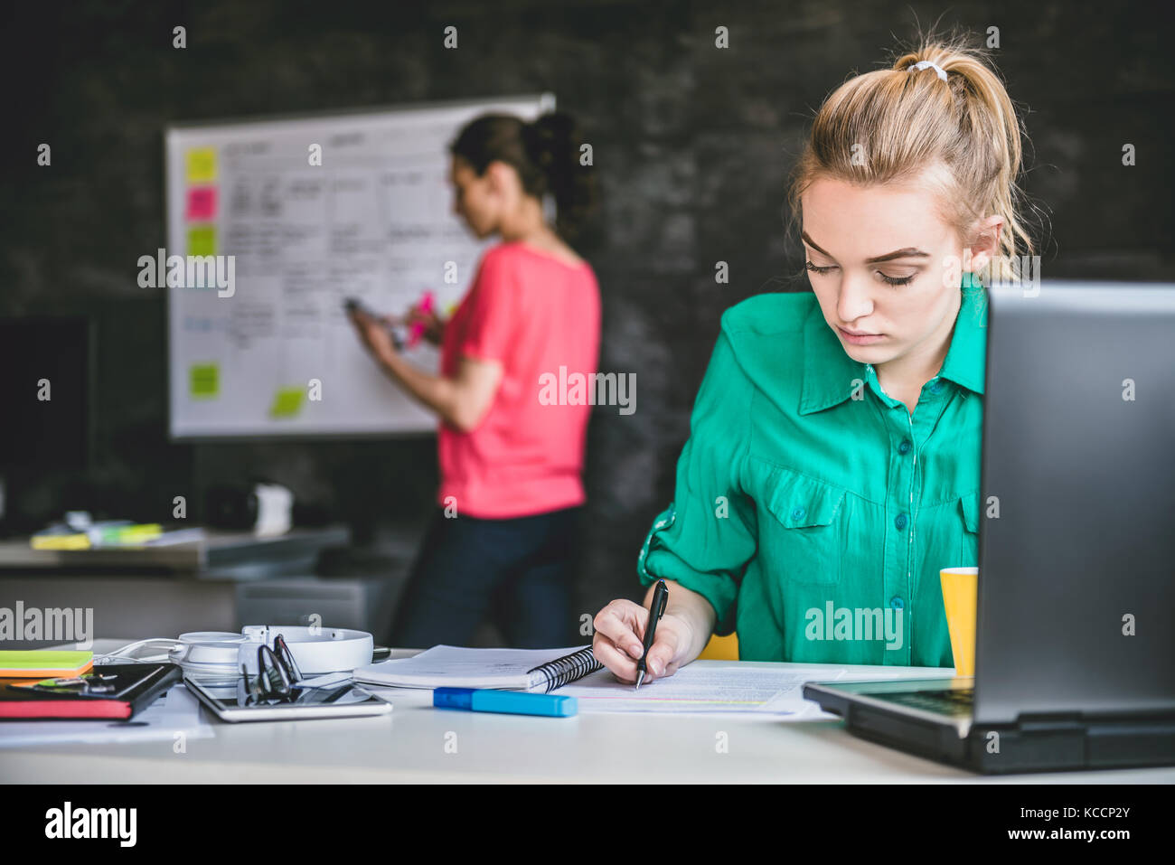 Young business woman wearing green shirt doing paperwork in the office and in the background is woman writing on whiteboard Stock Photo
