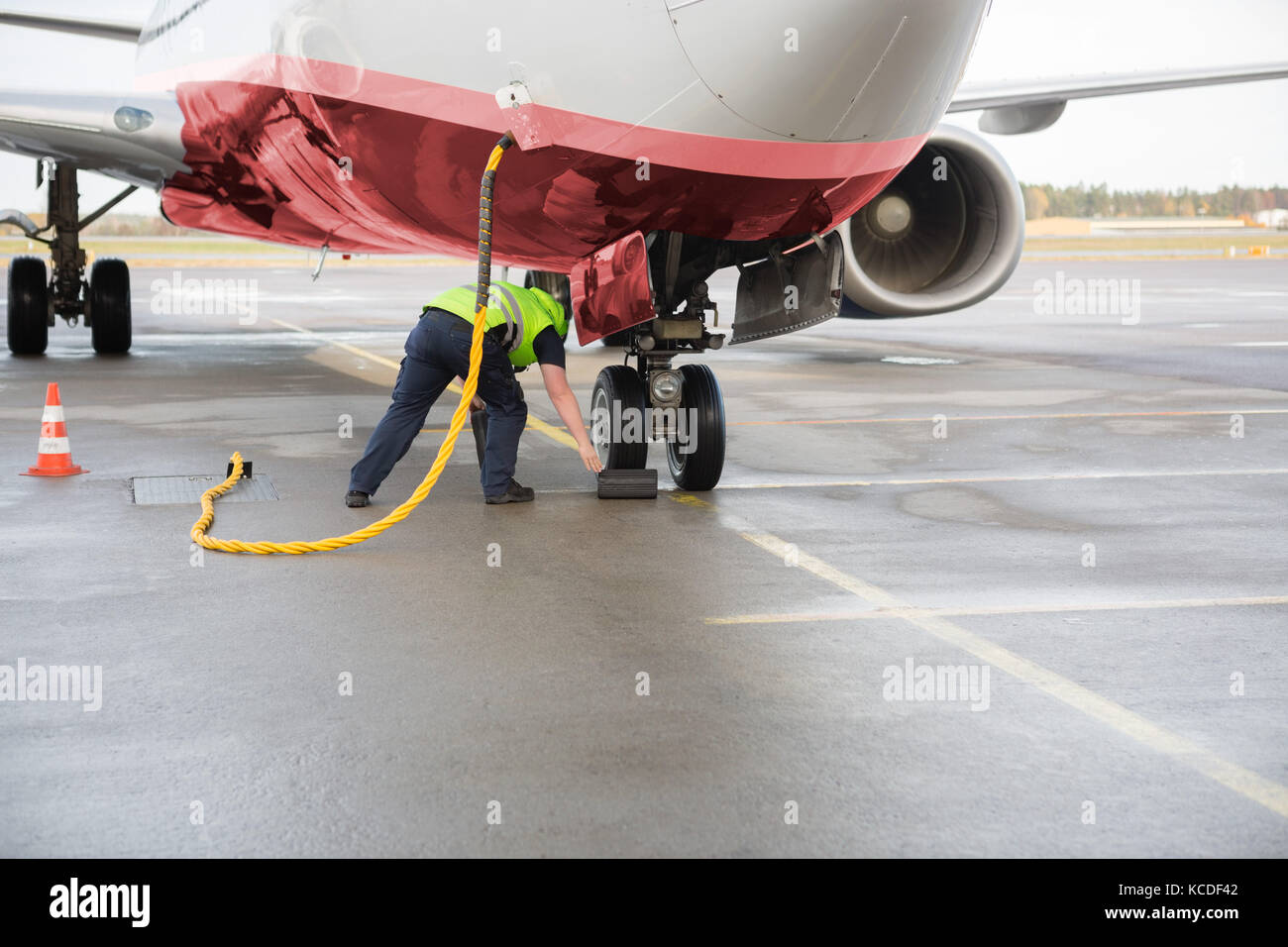 Full length of worker adjusting chock by airplane wheels while charging it on wet runway Stock Photo