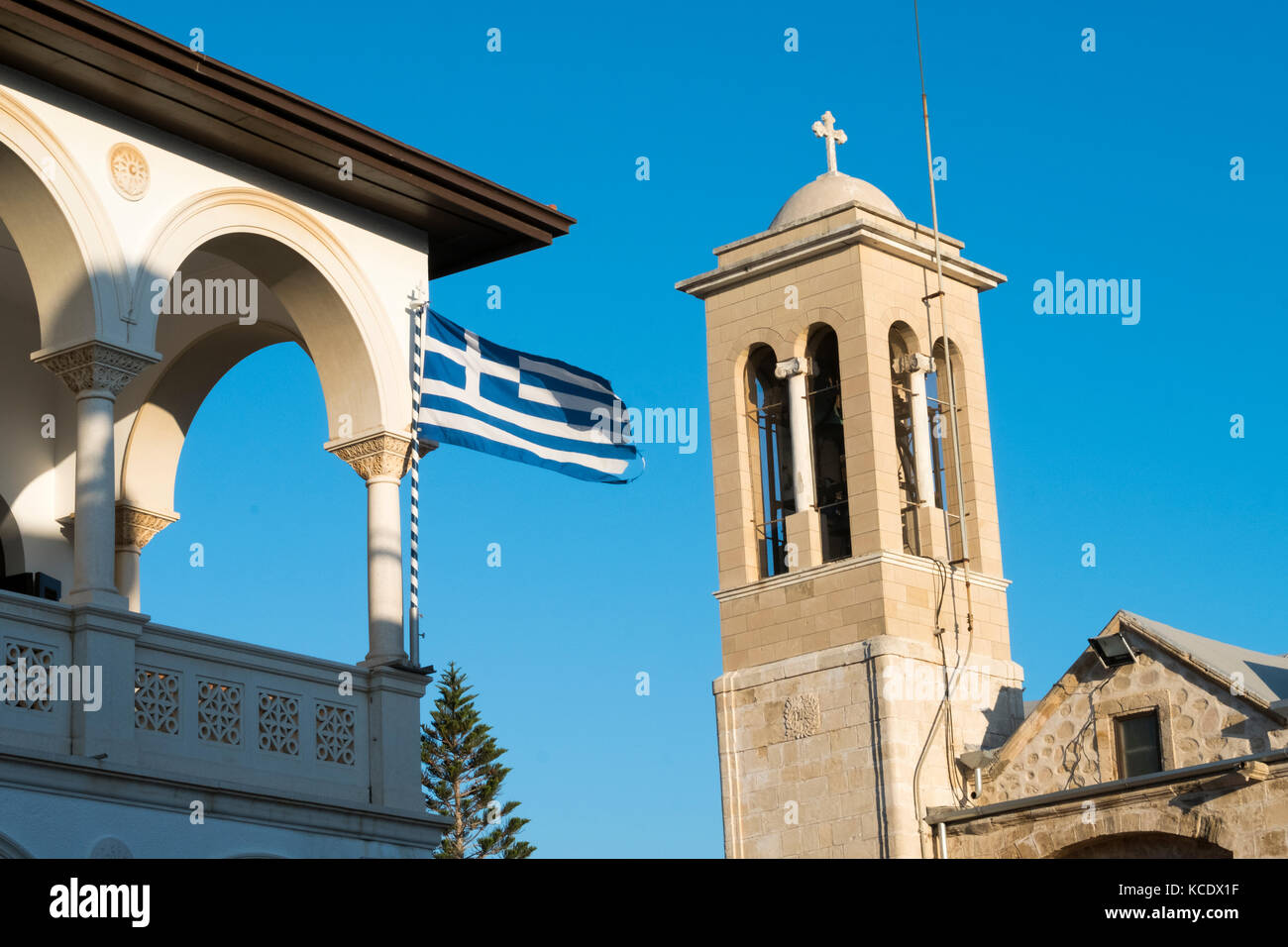 The flag of Greece flies on Holy Bishopric of Pafos, left and Agios Theodoros Cathedral on the right in Resistance Square ,Paphos Old Town, Cyprus Stock Photo