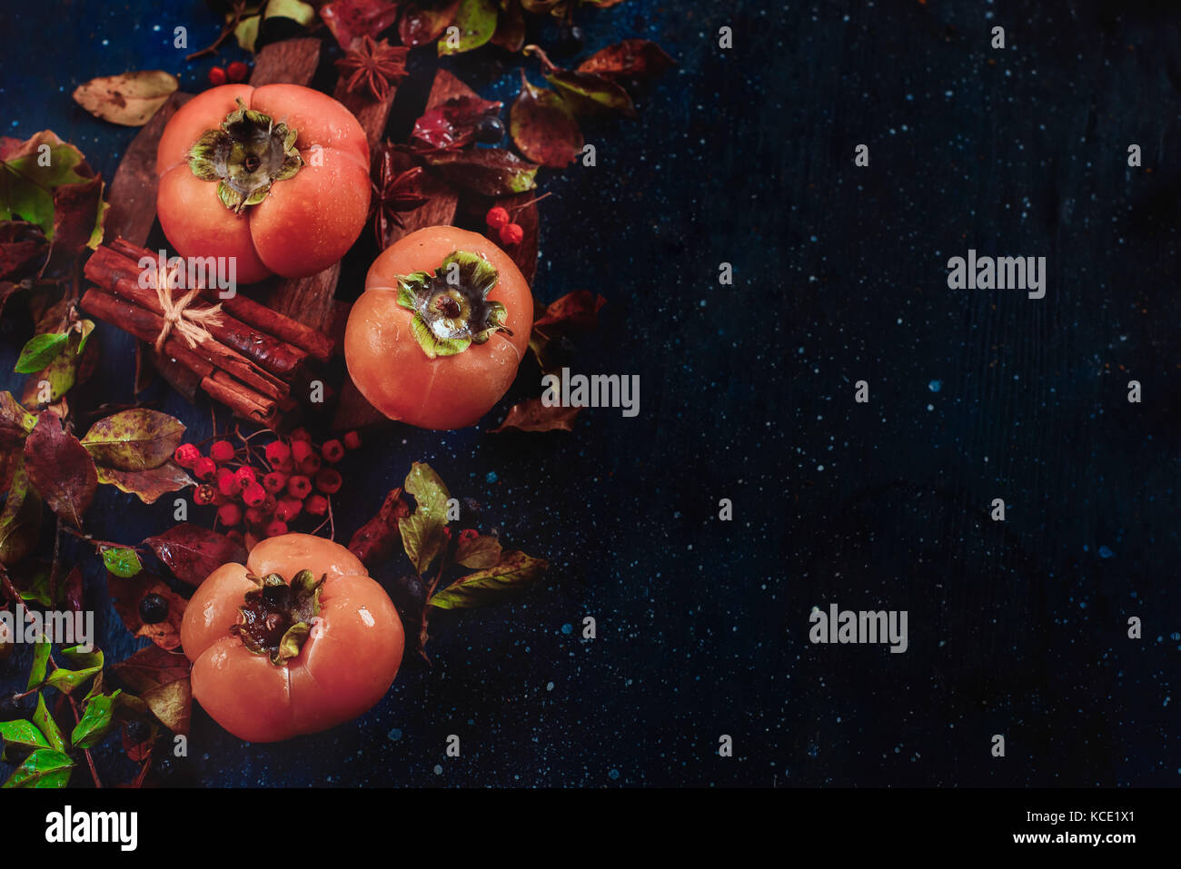 Ripe persimmons on a dark wooden background with autumn leaves, berries and cinnamon. Flat lay with copy space. Fruit in dark food photography. Stock Photo