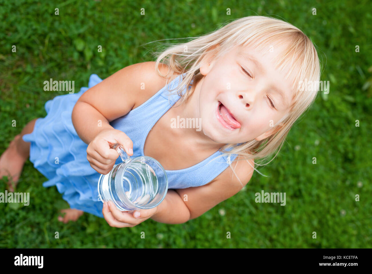 High angle view shot of blonde little girl wearing blue dress holding glass of water licking her lips with her eyes closed in a summer garden Stock Photo