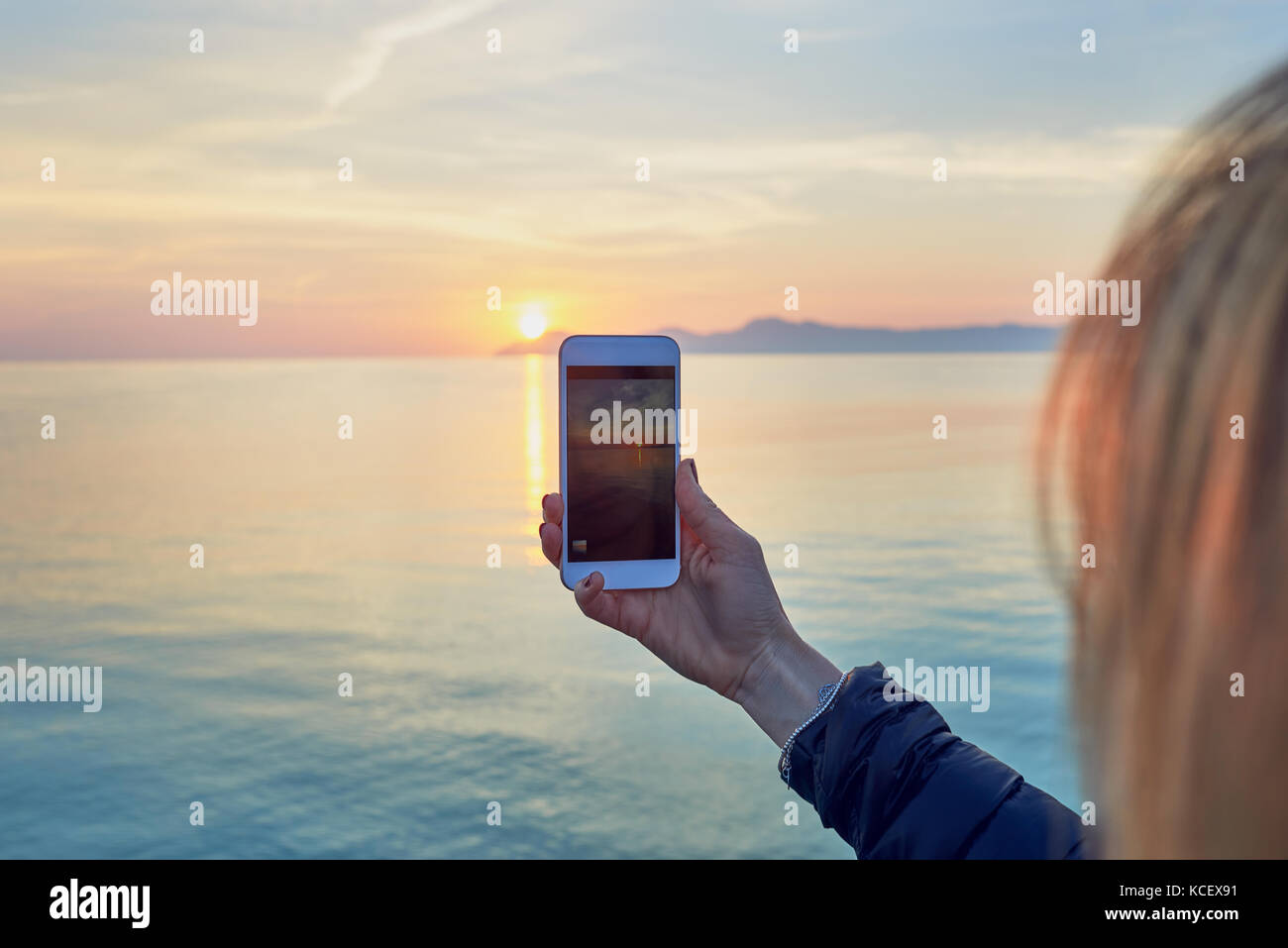Young blond woman taking a photo of a colorful ocean sunset on a mobile phone with a close up view of the screen of the cellphone with the sea and sky Stock Photo
