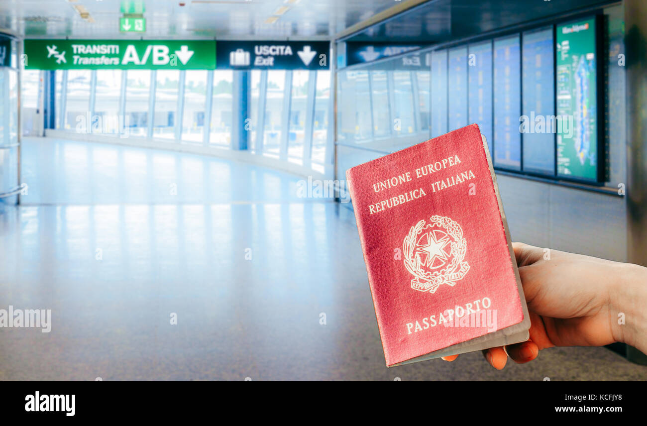 A hand holding an Italian passport at an Italian airport with flight information panels on the right Stock Photo