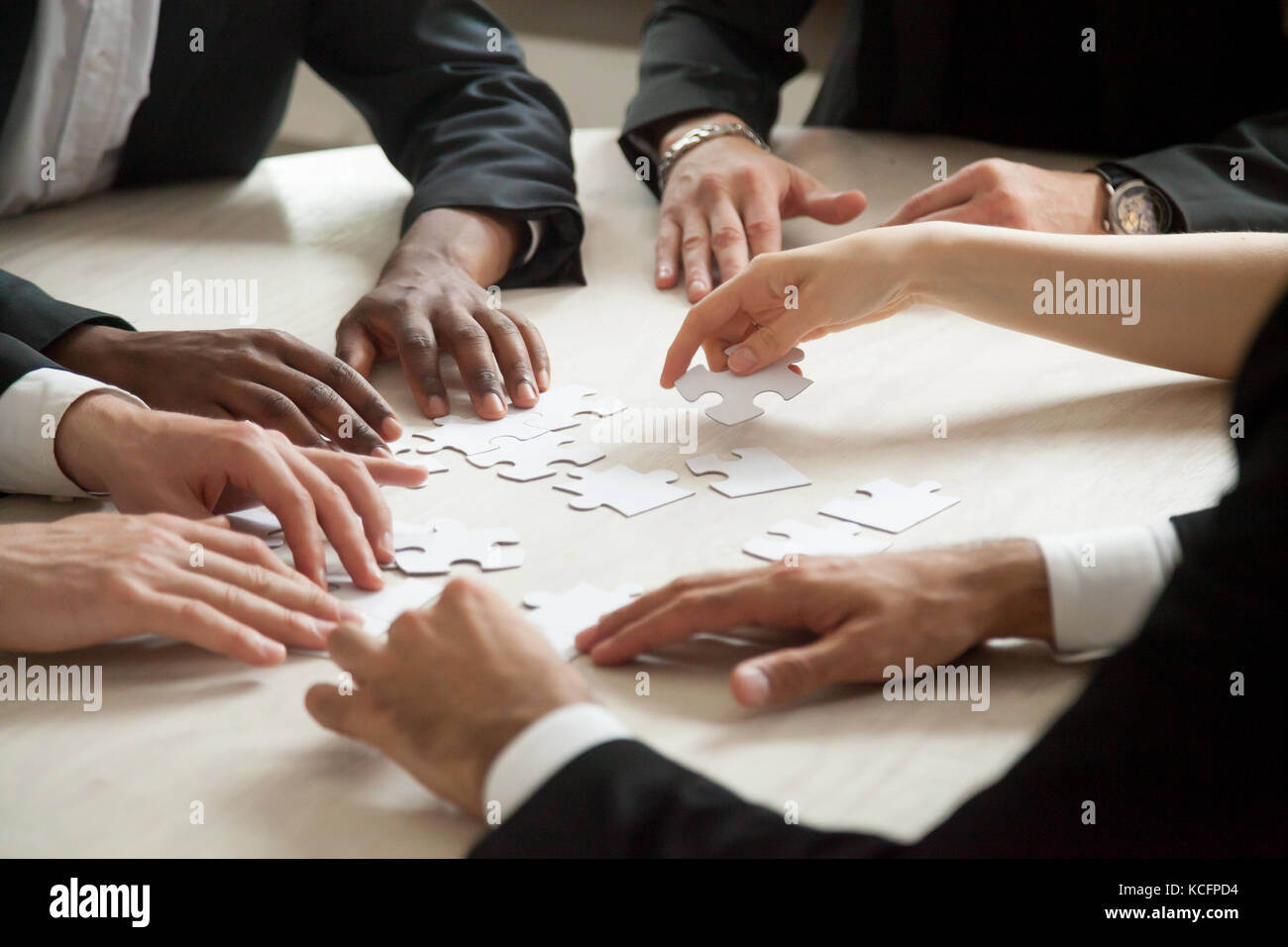 Close up of multiethnic team solving blank puzzle on table. Female hand offering puzzle piece, problem solution concept. Multi-racial executive employ Stock Photo