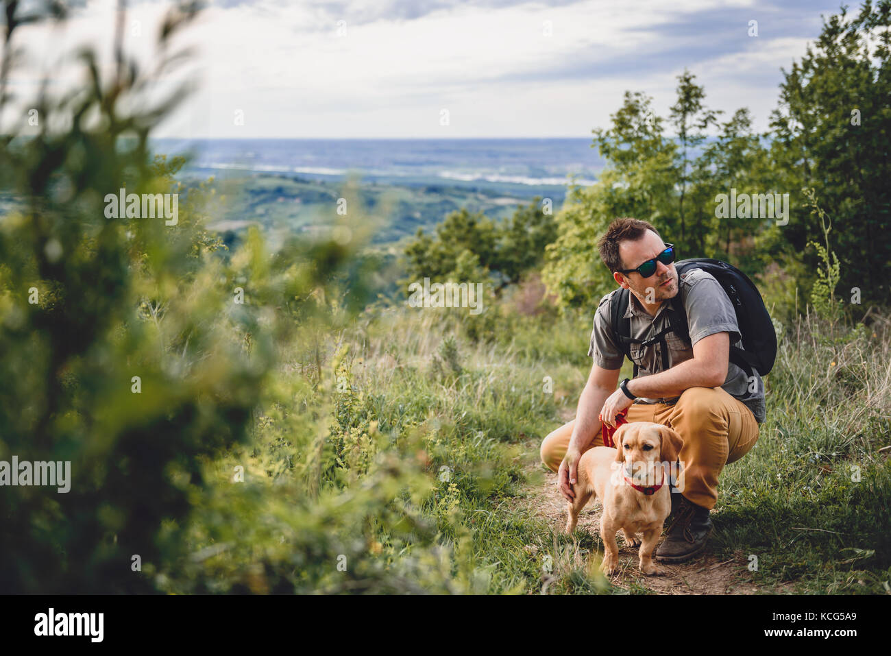 Man wearing sunglasses with a small yellow dog resting at the hiking trail Stock Photo
