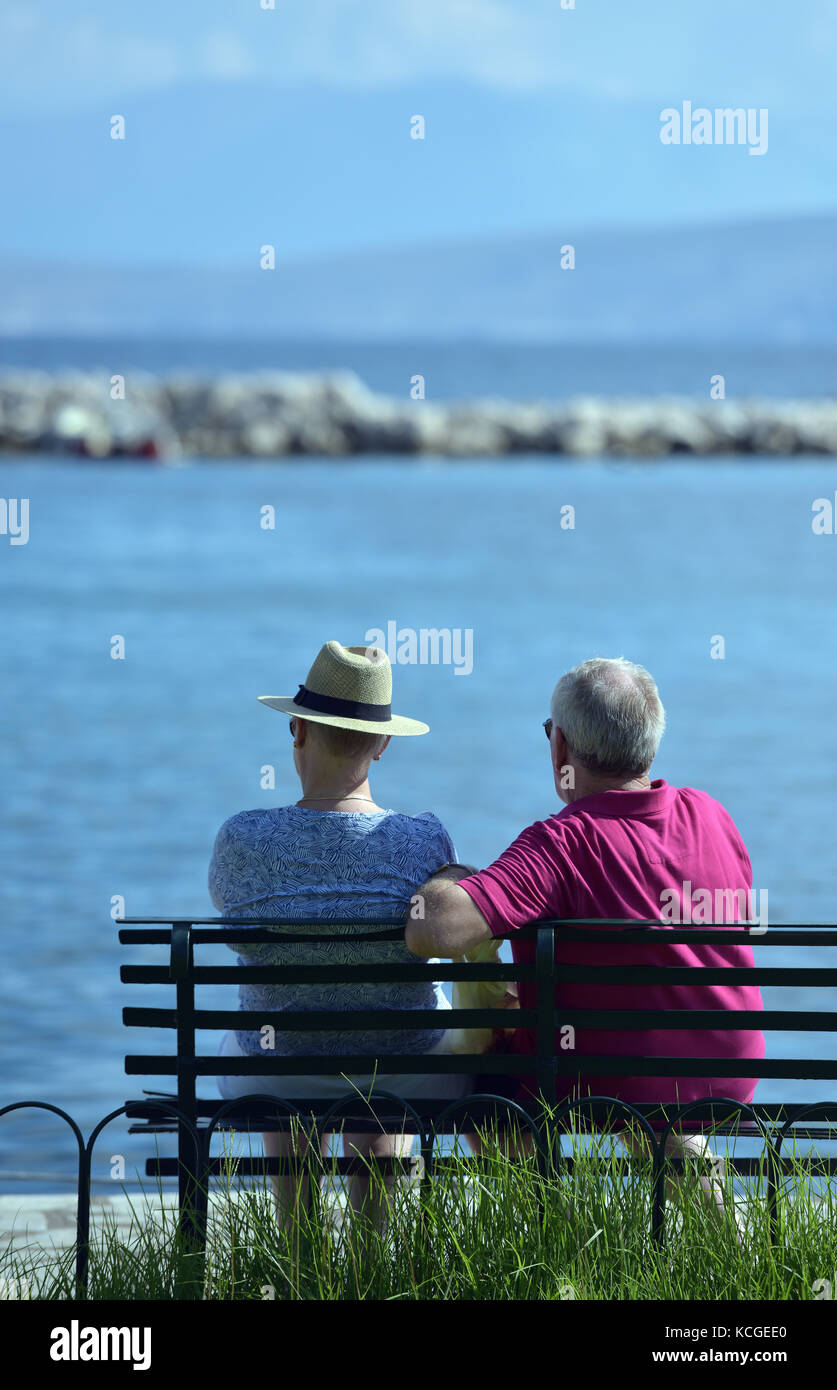 an older couple or a couple sitting on a bench looking out to sea with ...