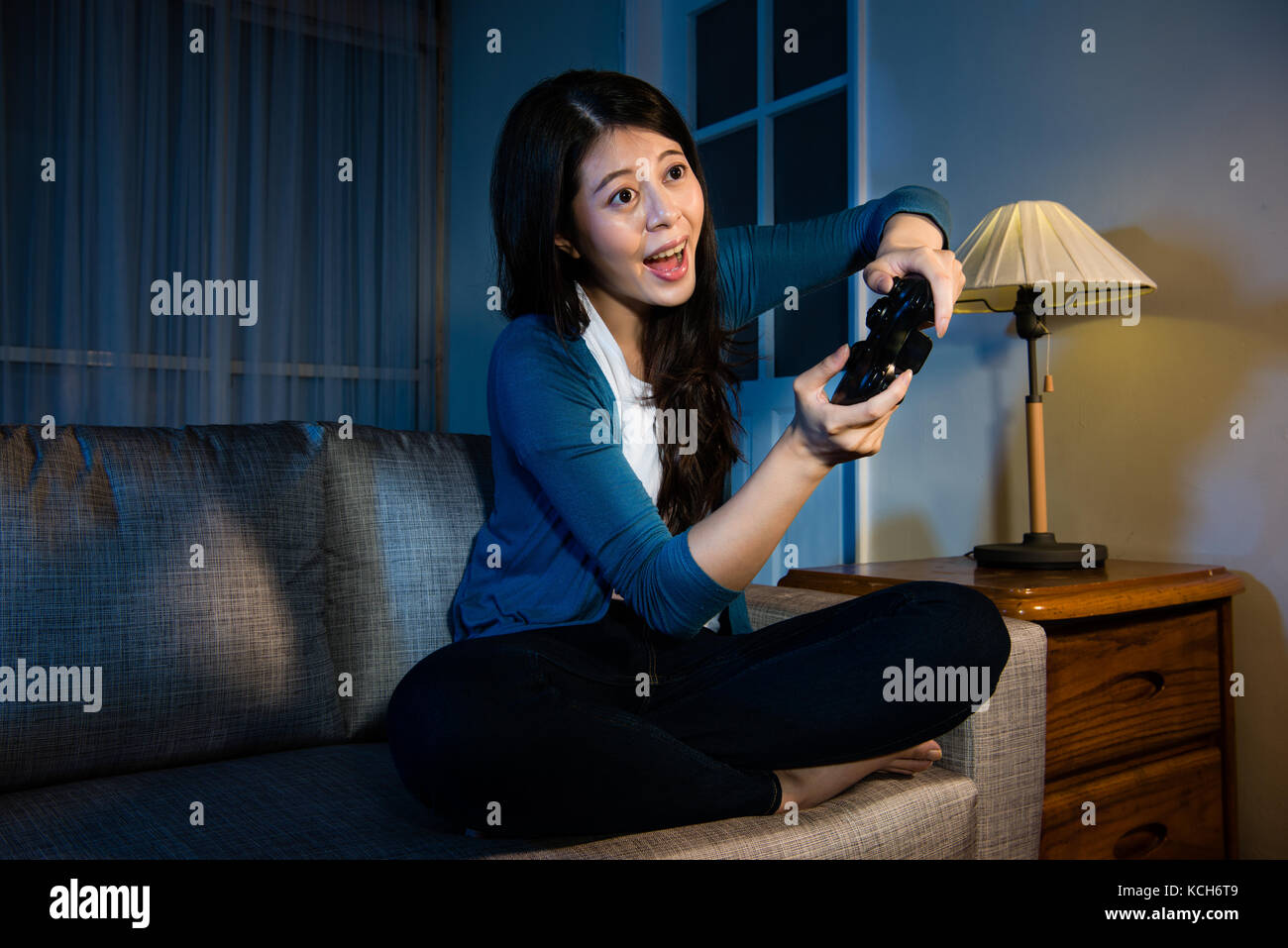pretty happy female student relaxing playing tv video game and holding controller with move body sitting in living room sofa couch at night. Stock Photo