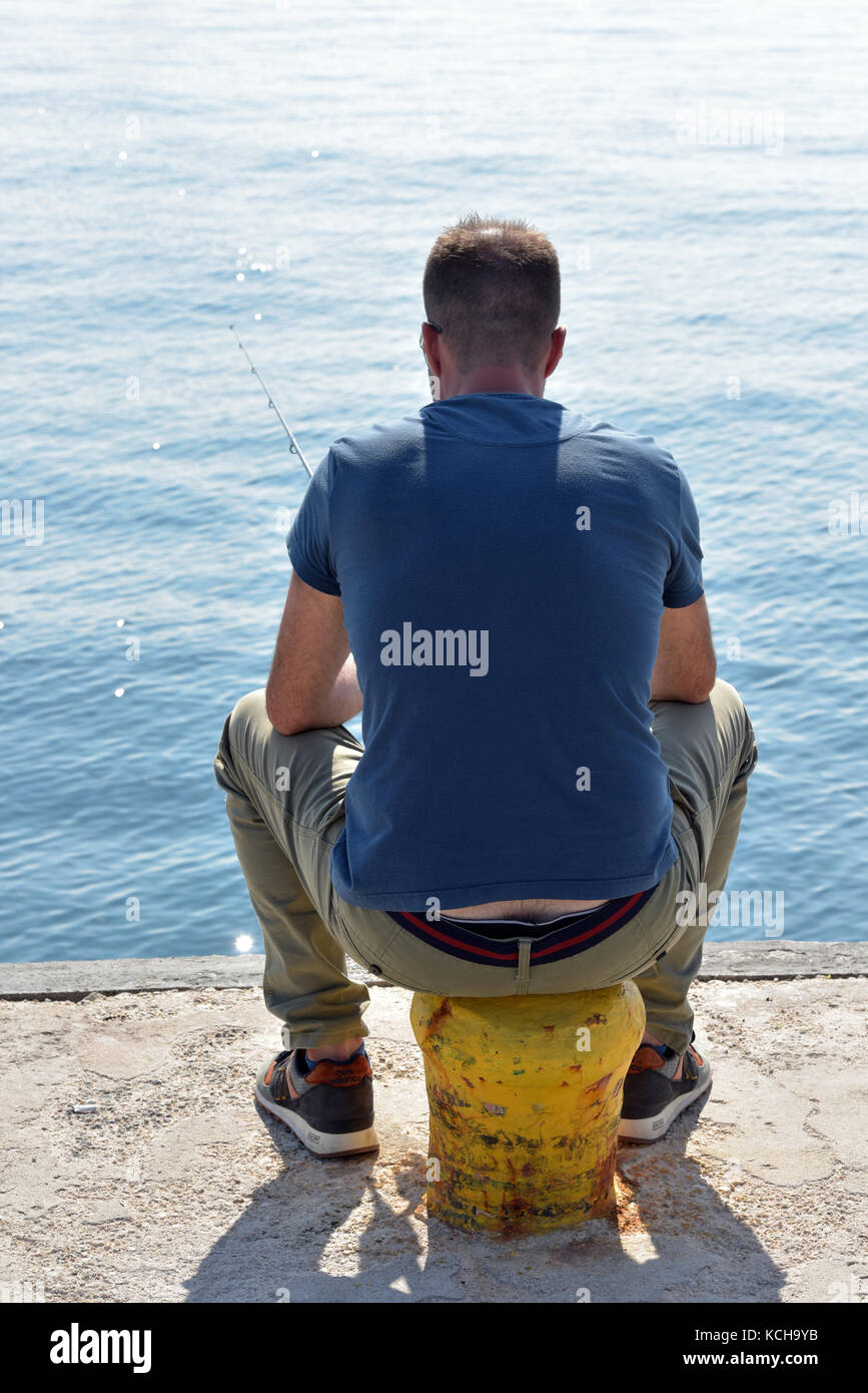 a man sitting on a bollard on the dockside or harbour wall fishing
