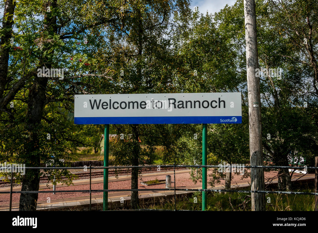 A white road sign with black letters saying Welcome to Rannach stands in front of trees partly masking the bright sunlit railway station Stock Photo