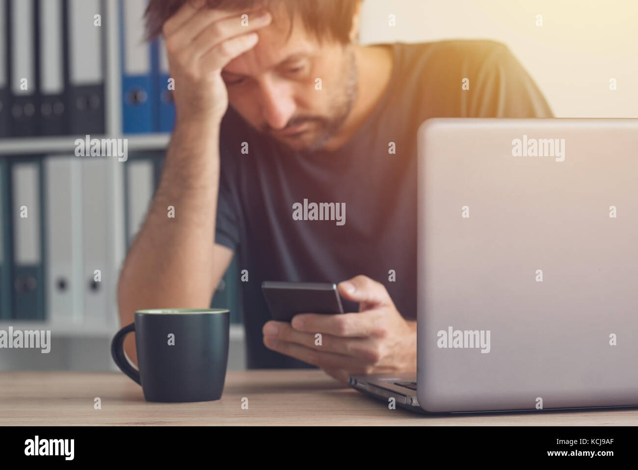 Worried freelancer reading bad news sms message on his smartphone in the office Stock Photo