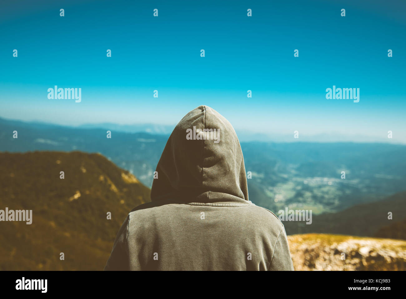 Unrecognizable female person standing at high mountain viewpoint. Rear view of young adult woman wearing hoodie looking at distance on horizon. Toned  Stock Photo