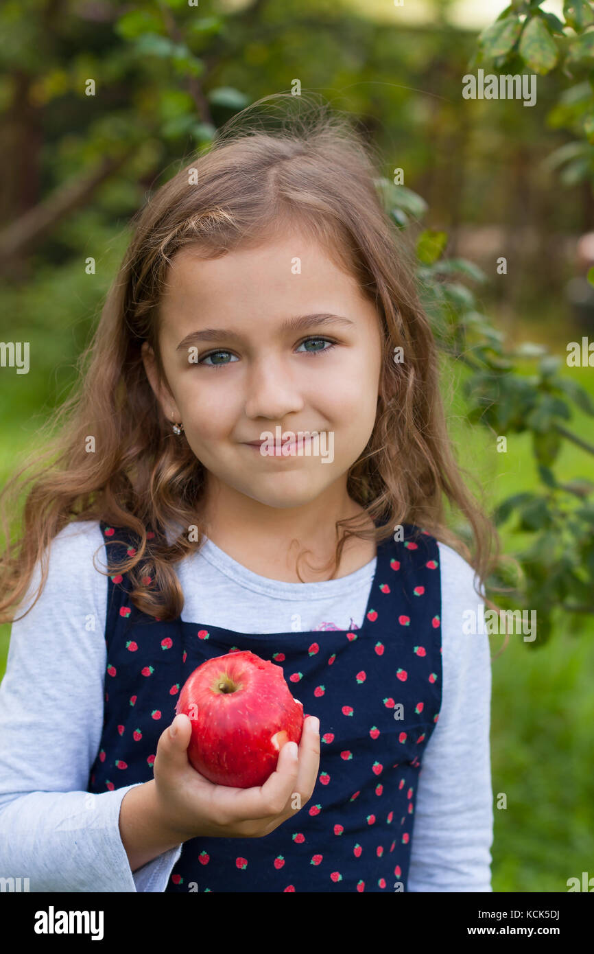 Cute Beautiful Russian Six-Year Girl Hold In Her Hand Fresh Ripe Tasty Red Eating An Apple, Smiling And Looking At Camera, Outdoor Close Up. Little Gi Stock Photo