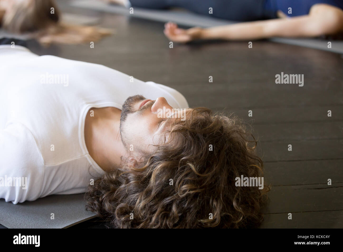 Close up of male head, young handsome man with curly hair, practicing yoga lesson with group in gym, lying in Dead Body exercise, doing Savasana, Corp Stock Photo