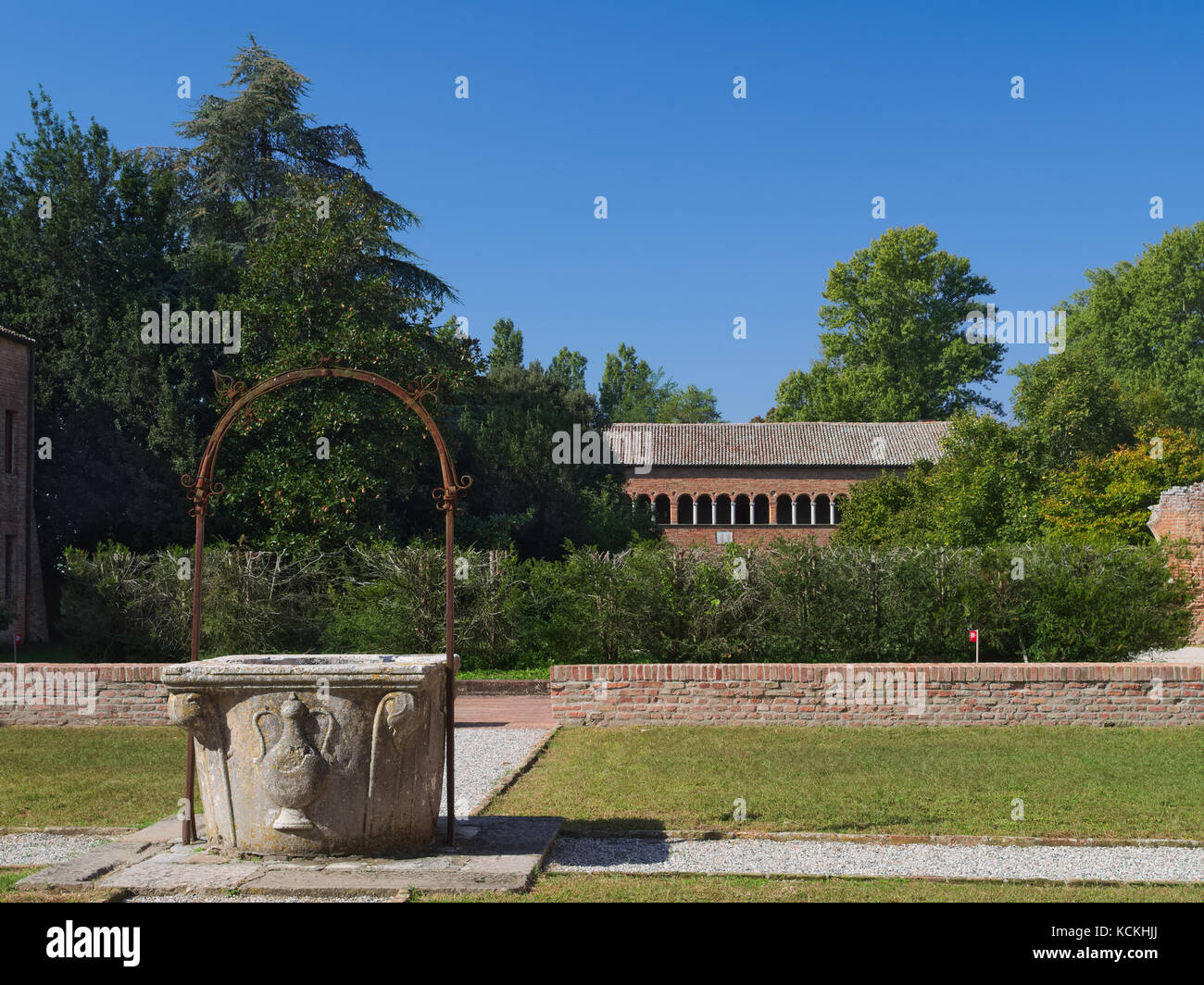 Historic well in the garden of the Pomposa Abbey Monastery with the “Palazzo della Ragione” building in the background. Codigoro, Ferrara, Italy Stock Photo