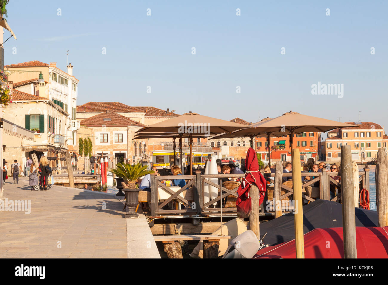 Murano, Venice,  Veneto Italy. Tourists enjoying sundowners canalside   at sunset in an open air restaurant, tavern  or trattoria on a deck over the w Stock Photo