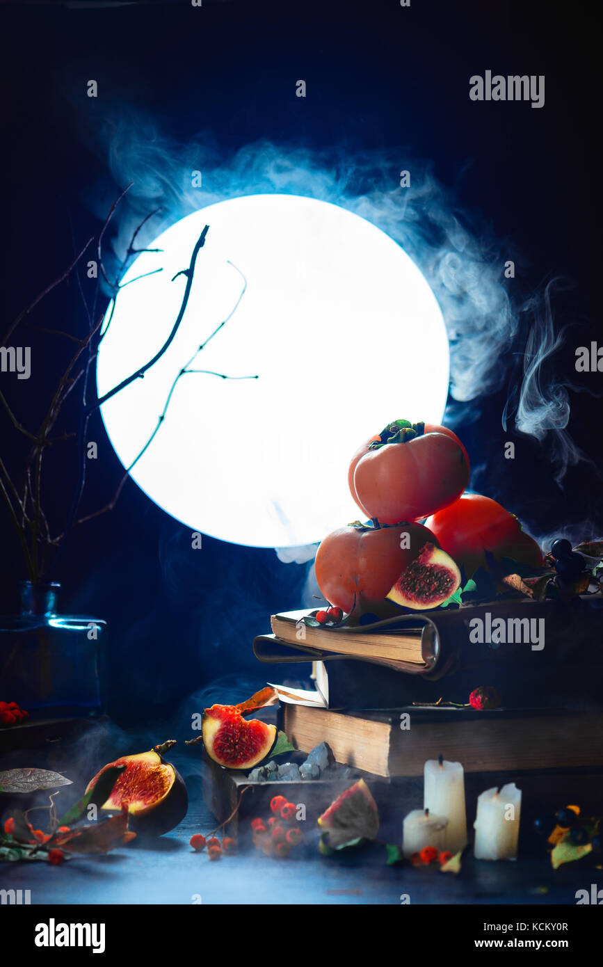 Harvest full moon still life with persimmon and figs on a stack of magical books. Dark Halloween concept with smoke. Magical equipment and candles. Stock Photo