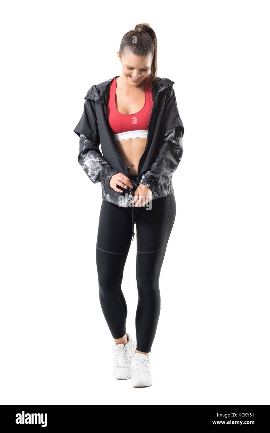 Smiling pretty sporty young woman getting dressed zipping tracksuit jacket. Full body length portrait isolated on white background. Stock Photo