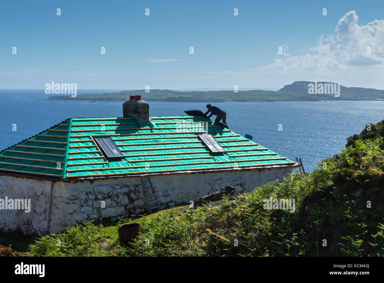 A worker restoring the roof of a cottage on the Isle of Eigg, Scotland, UK.  Behind is the Isle of Muck. Stock Photo