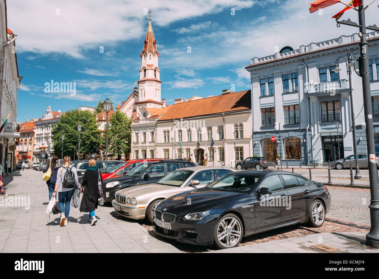 Vilnius, Lithuania - July 5, 2016: Young Women People Walking In Rotuses Street In Old Town. Parked Luxury Cars And St. Nicholas Church In Sunny Summe Stock Photo