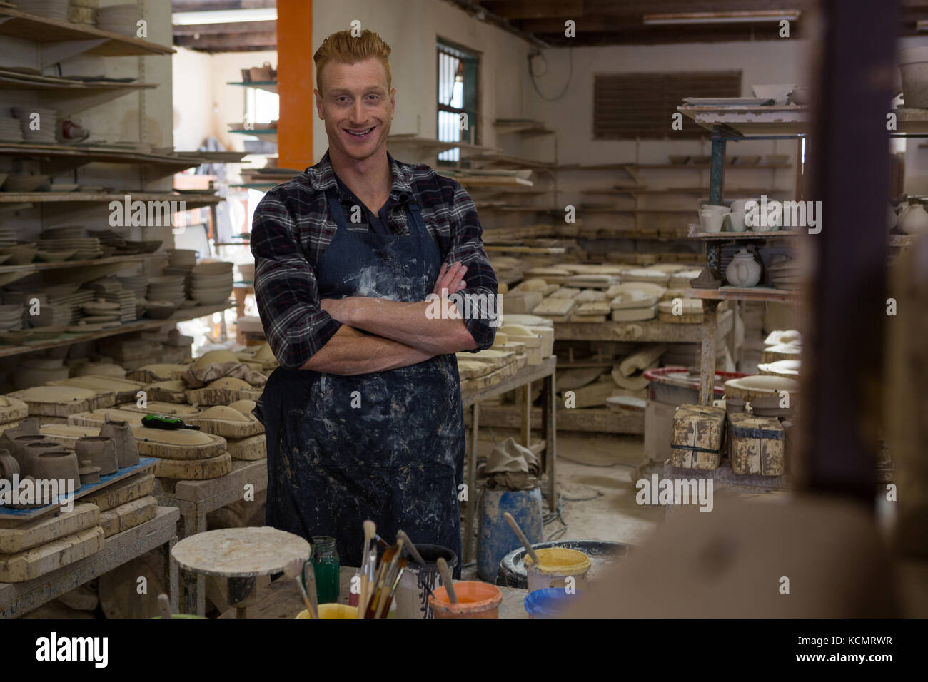 Portrait of male potter standing with arms crossed in pottery workshop ...