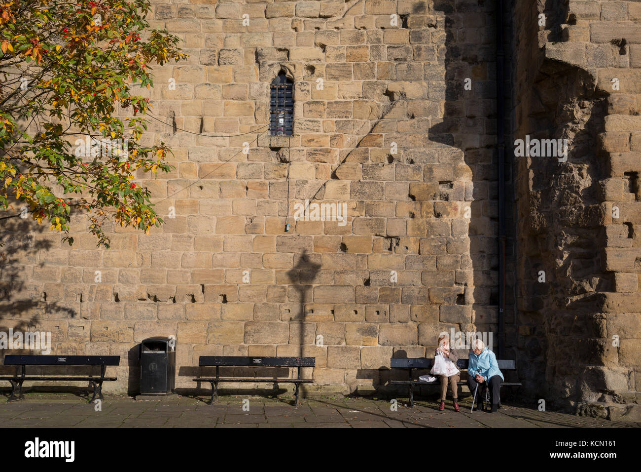 Locals sit in afternoon sunshine beneath the tall outer wall of Hexham's Moot Hall, on 29th September 2017, in Hexham, Northumberland, England. Originally, this gatehouse guarded the hall of the archbishops of York who were the Lords of the manor of Hexham for nearly 500 years until 1545. In later centuries the gatehouse became the setting for the Quarter Sessions of county magistrates and for the meetings of the town's Borough Courts, Since then it has been called the Moot Hall. Stock Photo