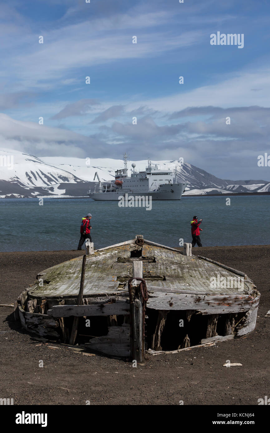 Adventure tourists walk near long abandoned water boats from the whaling era on a volcanic beach at Whalers Bay, Deception Island, South Shetland Islands Stock Photo